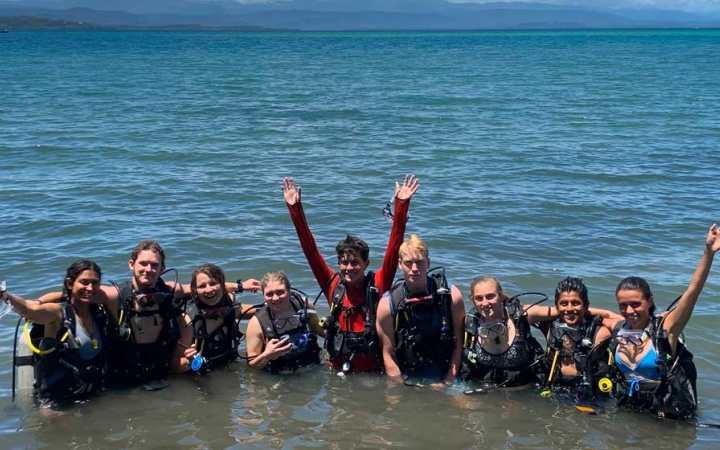 A group of students wearing Scuba gear stand in a line in shallow water and smile at the camera. A few of them raise their hands in apparent celebration. 